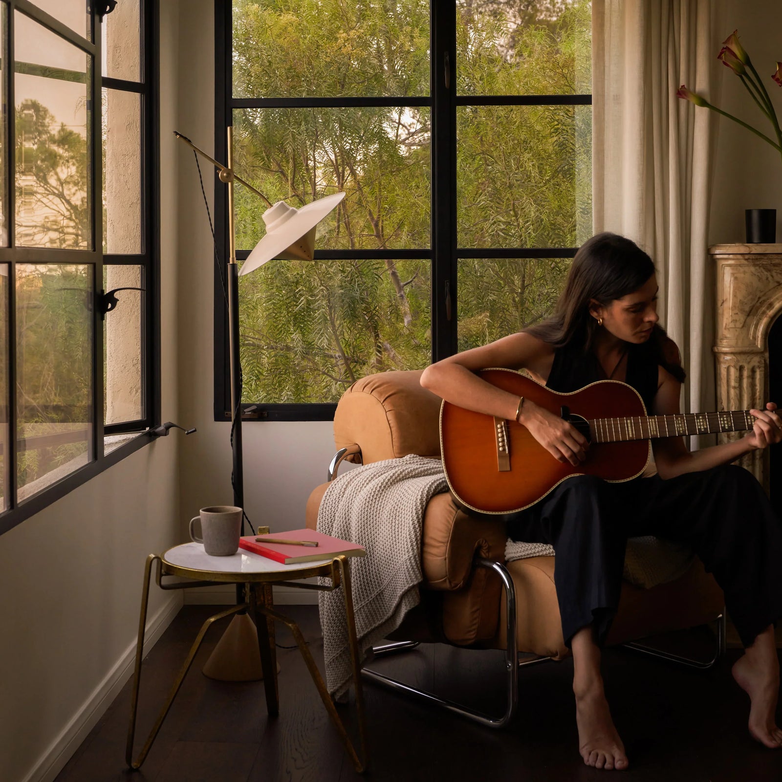 Woman playing guitar in a cozy room with large steel casement windows and a fireplace.