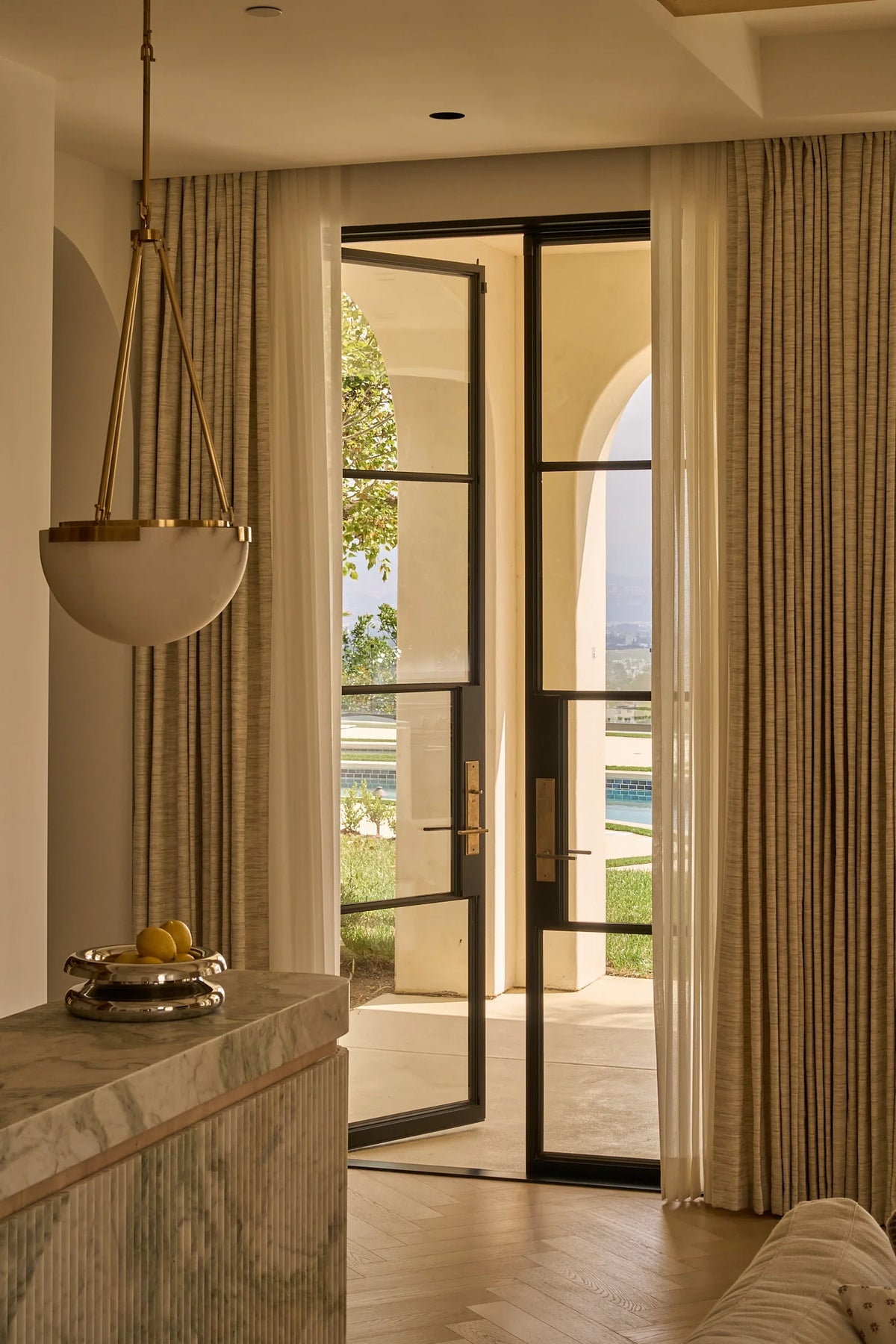 A luxury interior view featuring a pair of black-framed glass doors with beige drapes, looking out onto a patio and distant mountains. In the foreground, a marble console table with a hanging brass and glass pendant light is visible.