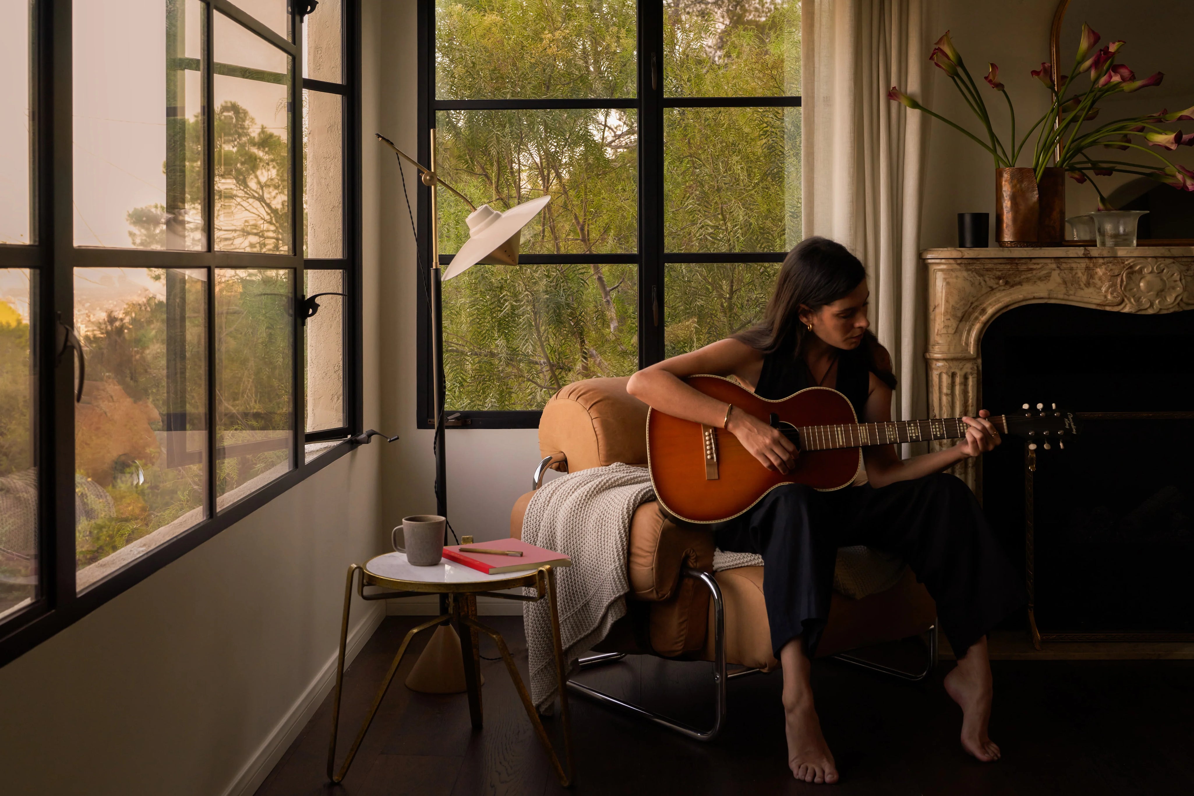 Woman playing guitar in a cozy room with large black casement windows and a fireplace.